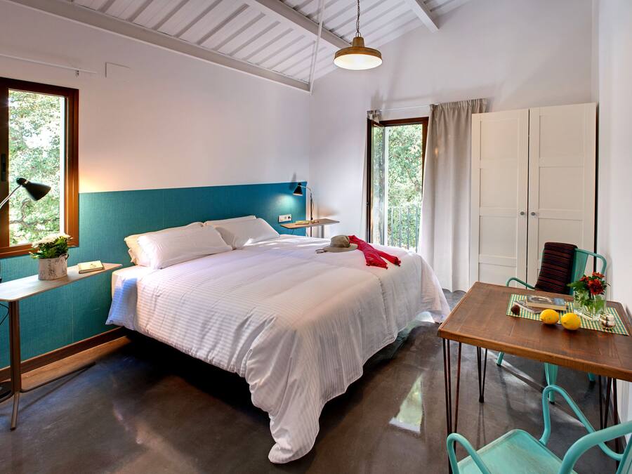 Un dormitorio con cama, mesita de noche, lámpara, ventana y una pequeña mesa con un florero y limones.