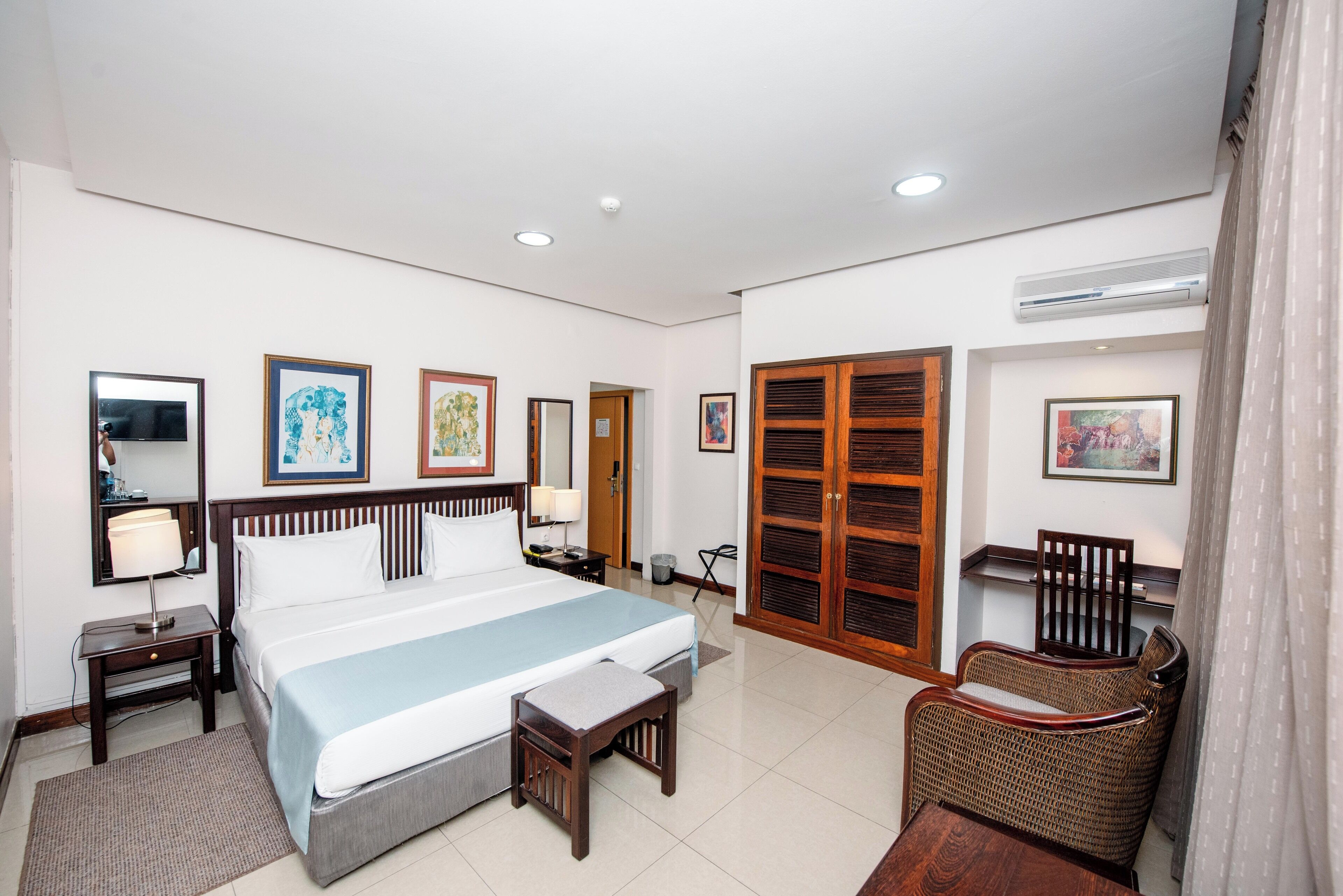 Una habitación de hotel con una cama grande, mesitas de noche, una silla, un armario y cuadros en las paredes.