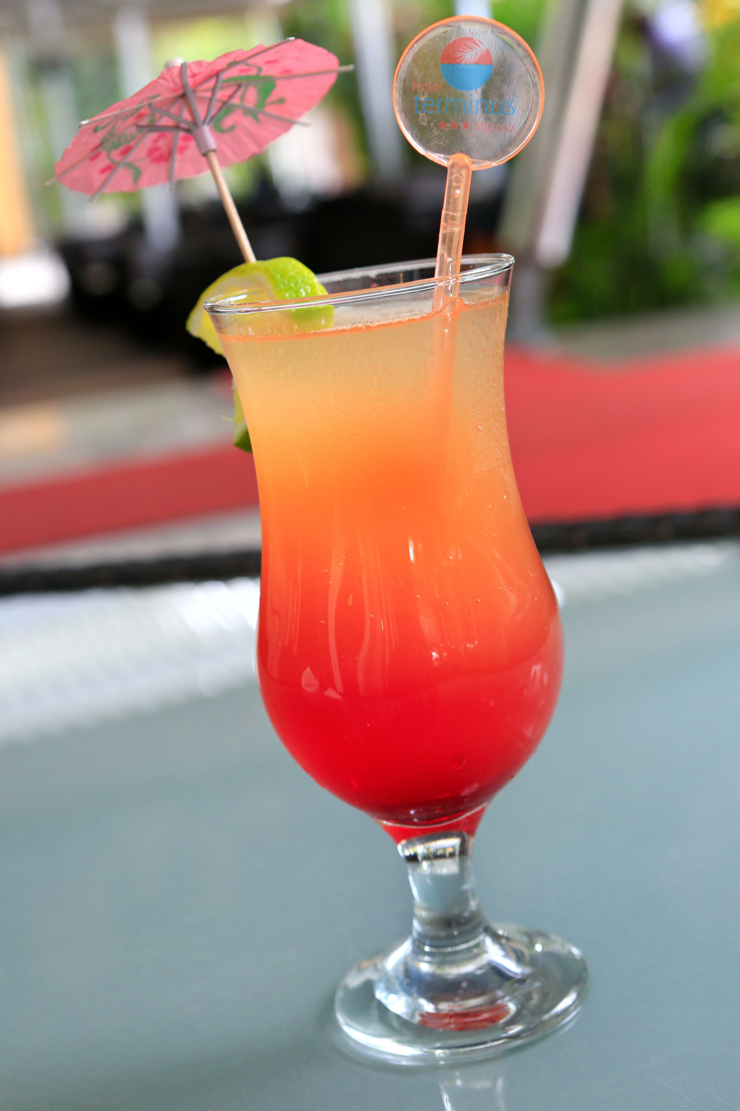 A cocktail in a tall glass with a lime wedge and a pink umbrella on a table.