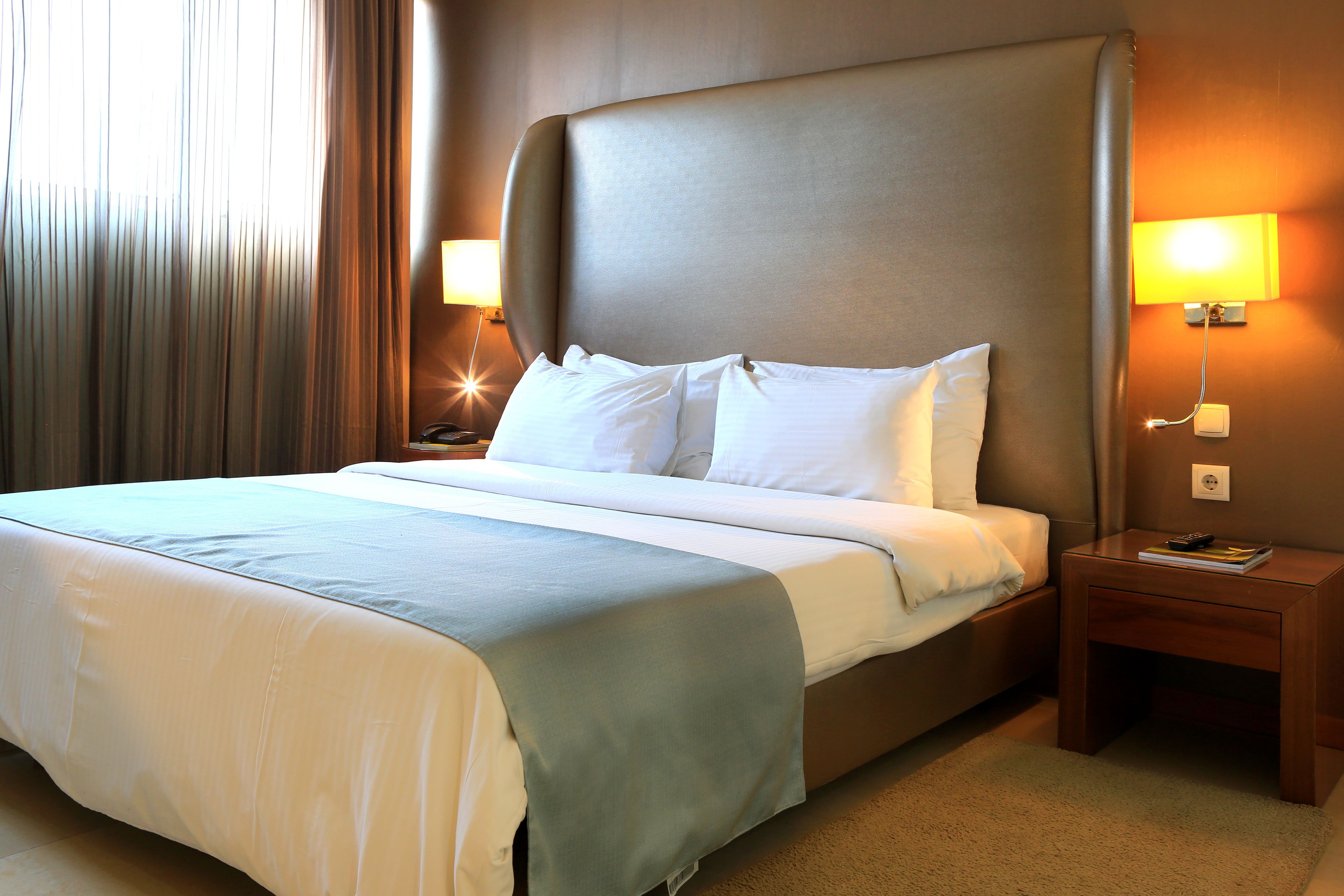 A neatly made bed with white linens and a light blue accent, a wooden nightstand with a lamp, and a wall-mounted light fixture.