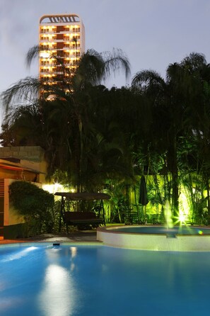 Una piscina rodeada de abundante vegetación y un alto edificio de hotel al fondo.