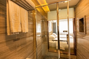 A modern hotel bathroom with a glass-enclosed shower, a towel rack with towels, and a bed visible through the glass.