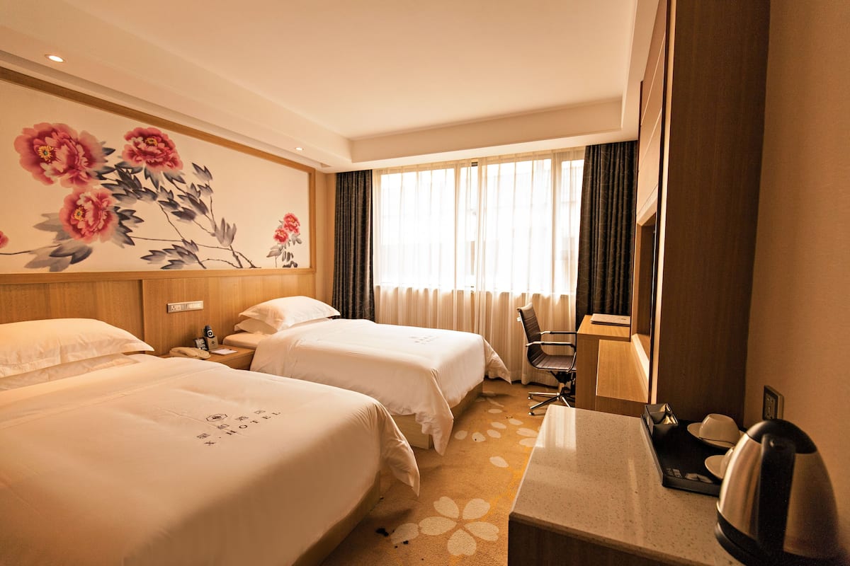 A hotel room with two beds, a desk, a chair, a coffee maker, and a large window with curtains.