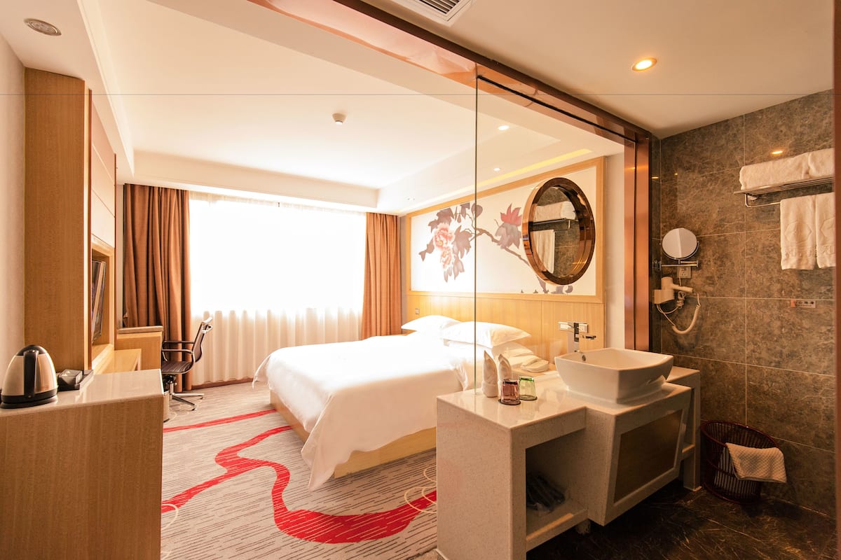 A hotel room with a bed, bedside table, mirror, and a bathroom with a sink and shower.