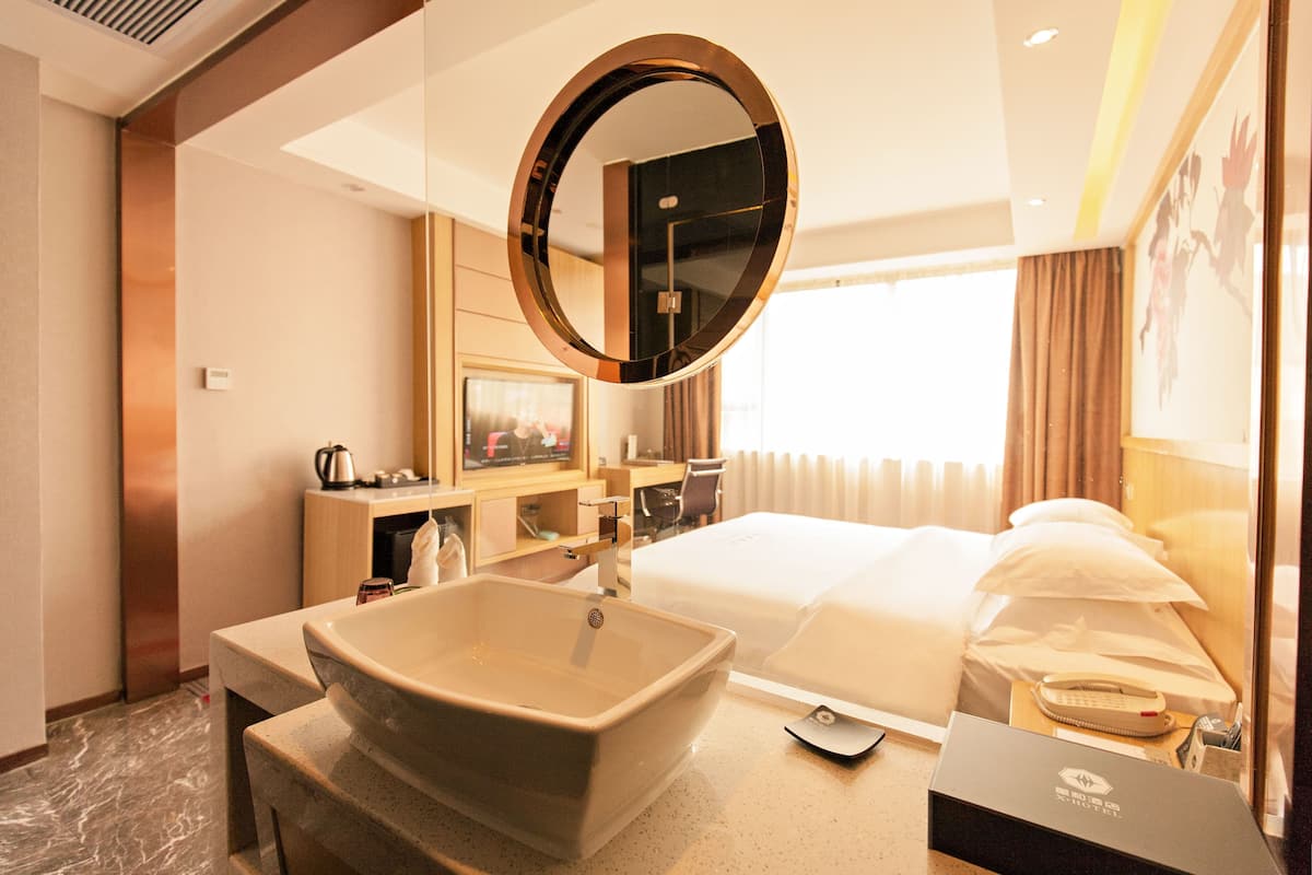 A hotel room with a large oval sink, a round mirror, a bed with white linens, a bedside table with a phone and a lamp, and a window with curtains.