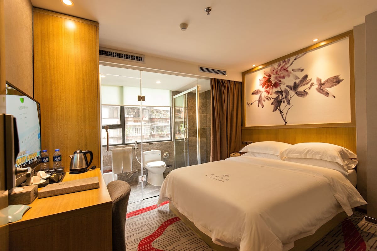 A hotel room with a bed, a desk with a computer, a coffee maker, and a bathroom visible through a glass door.