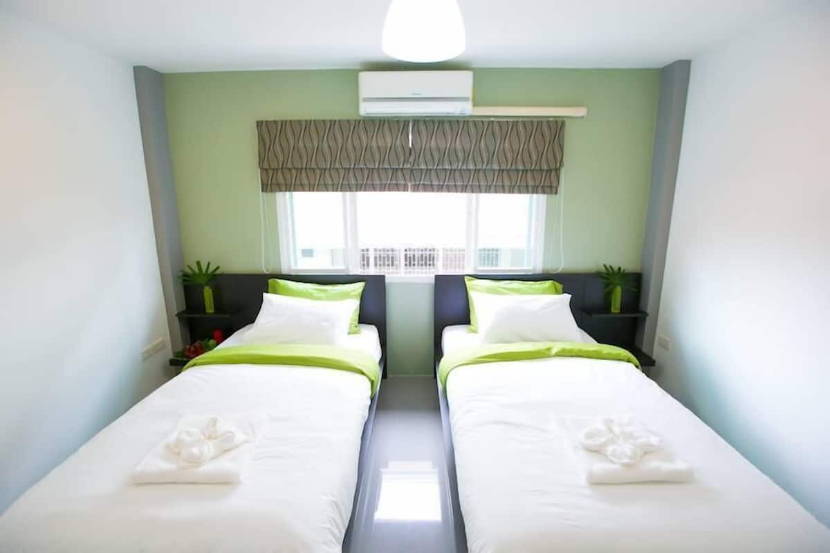A hotel room with two single beds, a window with a patterned valance, an air conditioning unit, and bedside plants.