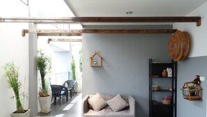 A modern living room with a grey sofa, wooden beams, a small house-shaped decoration, and a black shelving unit with baskets. - White Monkey Guesthouse (Phetchaburi)