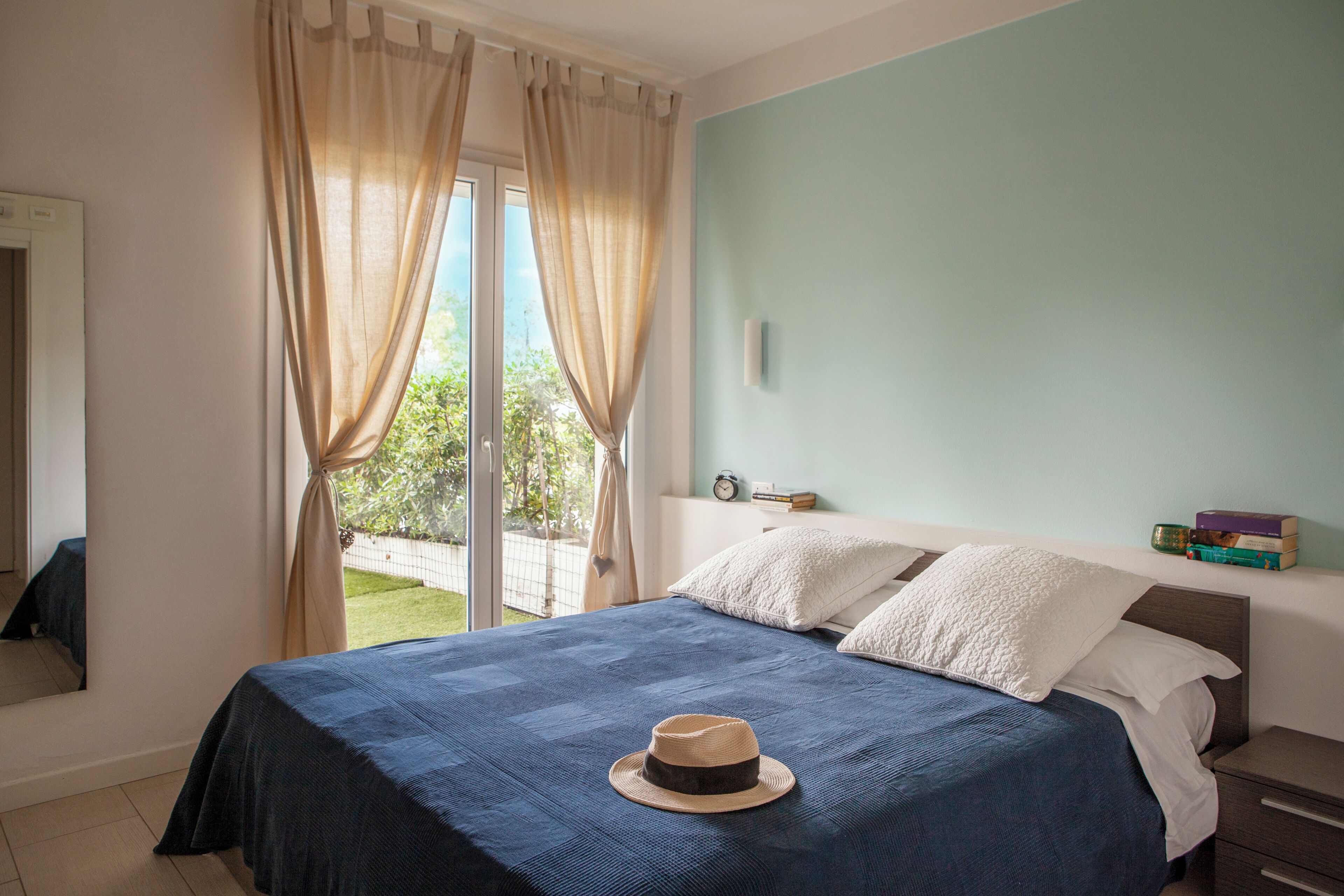 Une chambre à coucher avec un lit, un chapeau posé sur le lit, une fenêtre avec des rideaux et une vue sur la verdure à l’extérieur.