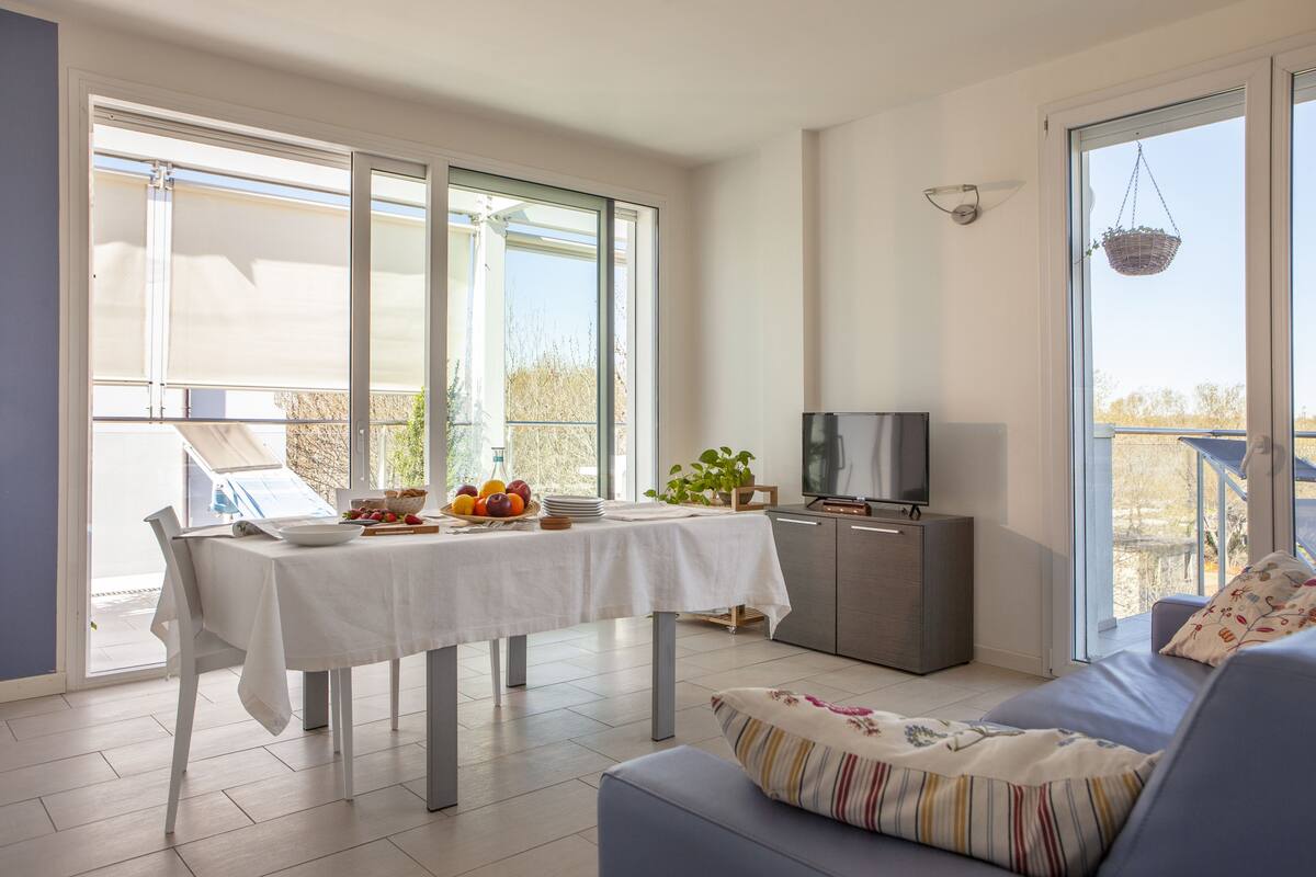 Un salon moderne comprenant un coin repas, de grandes fenêtres, un téléviseur à écran plat et un balcon agrémenté d’un pot à suspendre.