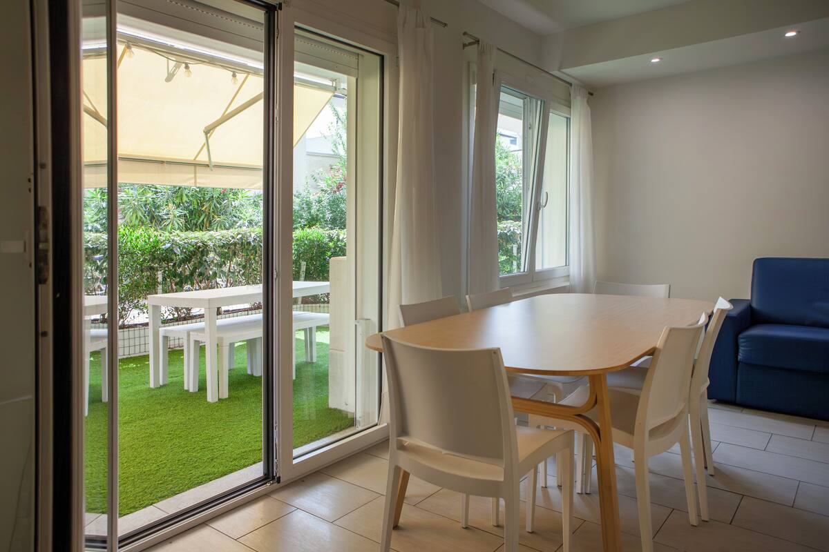 Une pièce comprenant une table à manger, des chaises, un canapé et une porte-fenêtre coulissante donnant sur un espace extérieur avec une terrasse et de la verdure.