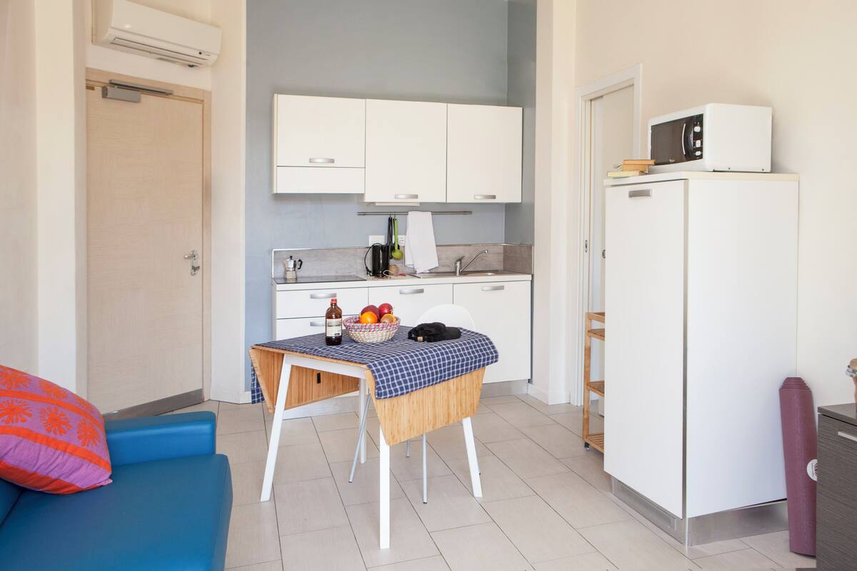 Une cuisine compacte avec des meubles blancs, un four à micro-ondes, une petite table recouverte d’une nappe à carreaux et un canapé bleu agrémenté d’un coussin rouge.