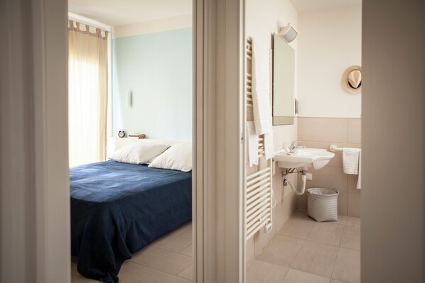 A bedroom with a bed, a bedside table, a window with curtains, and a bathroom with a sink, mirror, and towel rack.
