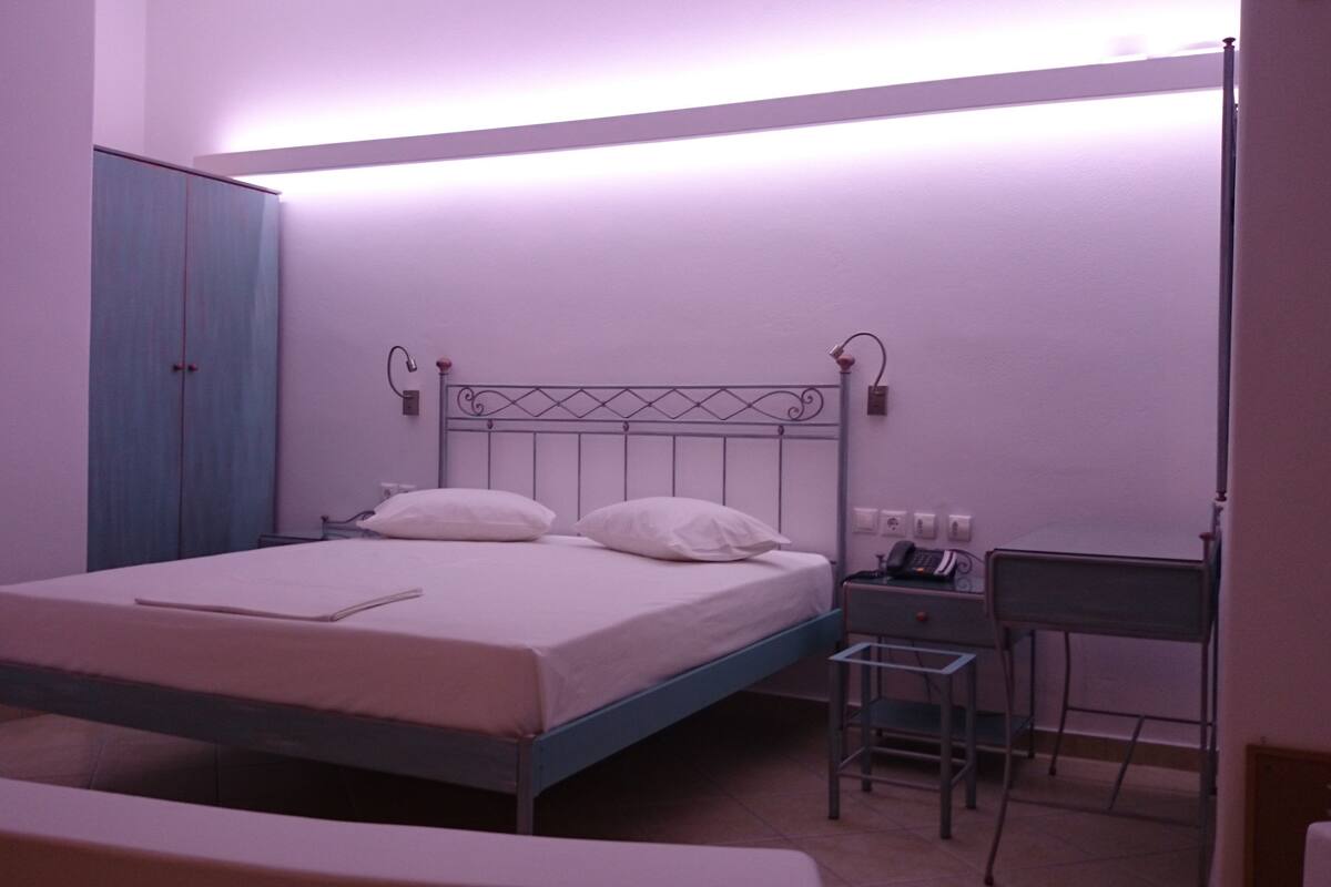 A single-bed room with a metal headboard, a bedside table with a lamp, a chair, and a wardrobe.