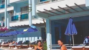 A swimming pool with lounge chairs and umbrellas, a multi-story building with balconies, and a clear sky.