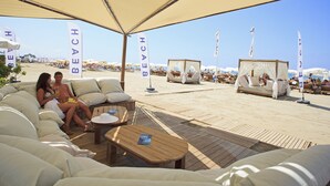 Uma área de estar à beira-mar com assentos acolchoados e guarda-sóis, um deck de madeira e bandeiras que indicam um ambiente de resort de praia.