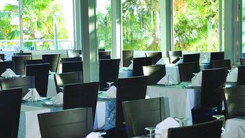 Restaurante com mesas postas para refeições, grandes janelas com vista para o verde e um lustre moderno.