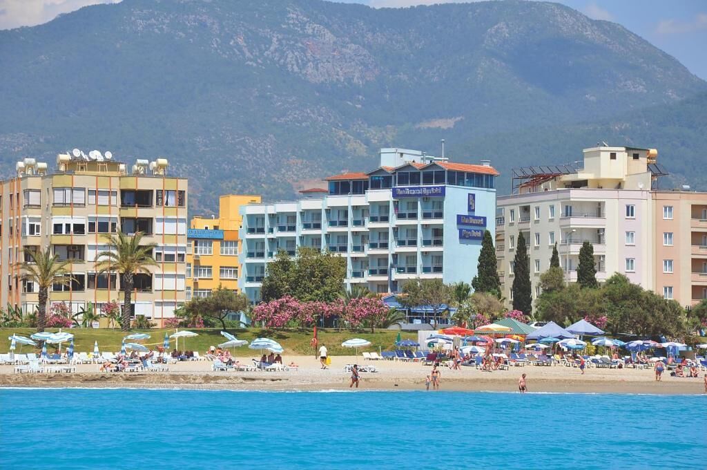 A coastal hotel with multiple floors, a beach in the foreground, and mountains in the background.