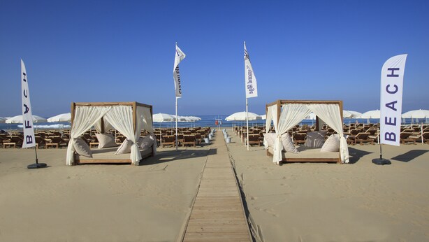 Um resort à beira-mar com duas camas de praia cobertas por toldos, um deck de madeira e bandeiras com a palavra 'BEACH'.