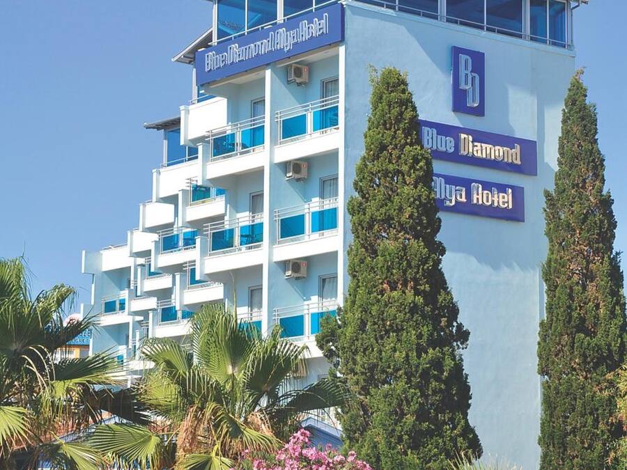 Een meerhoog hotel met balkons, een opvallend bord met 'Blue Diamond Myra Hotel' en omliggende begroeiing, waaronder palmbomen en bloeiende planten.