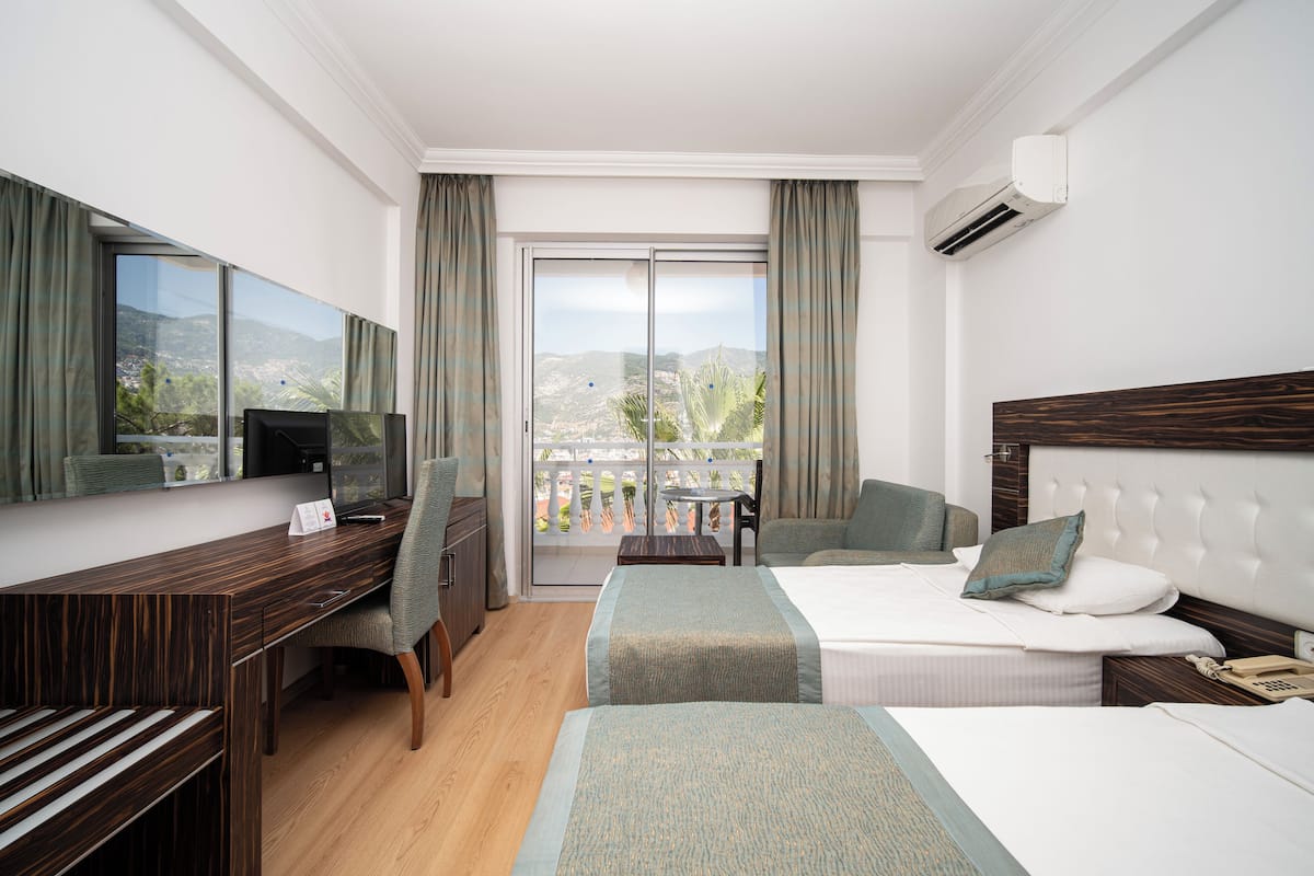 A hotel room with a wooden desk, two beds, a TV, and a balcony with a view of the mountains.