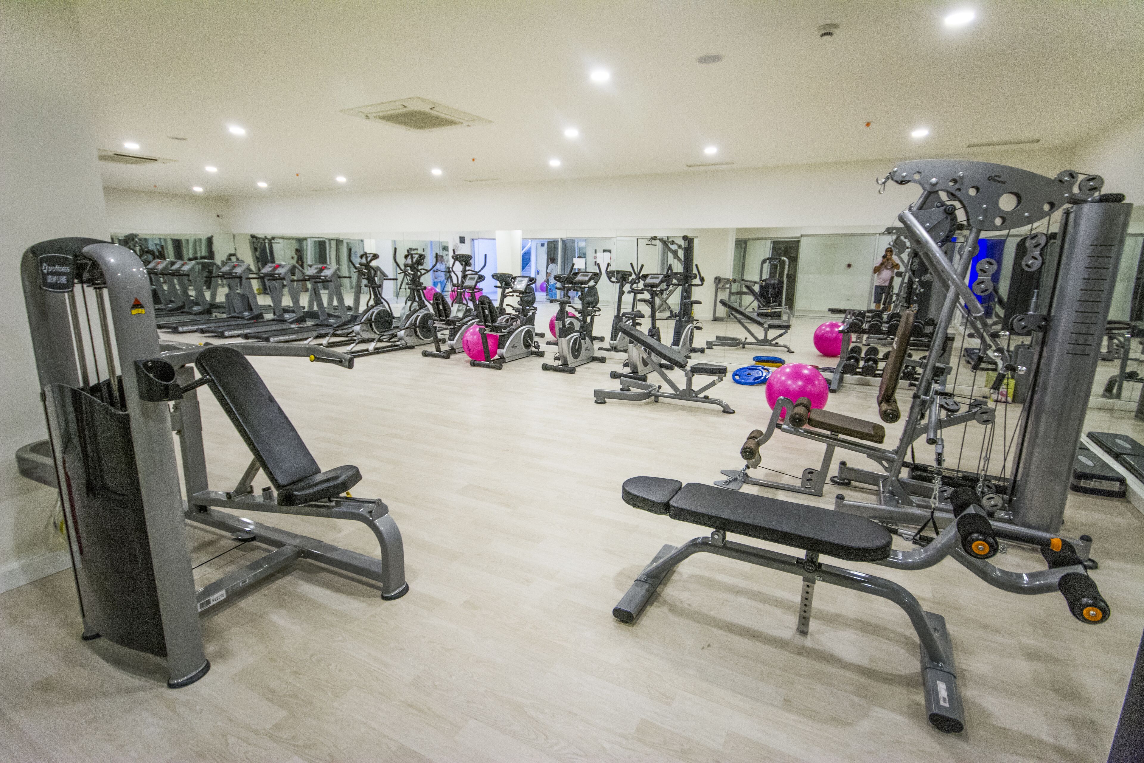 A gym with various exercise machines, mirrors, and a person in the background.