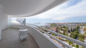 Un balcon doté d’une balustrade blanche, d’une table et de chaises, offrant une vue sur un paysage urbain côtier avec des bâtiments et une étendue d’eau.