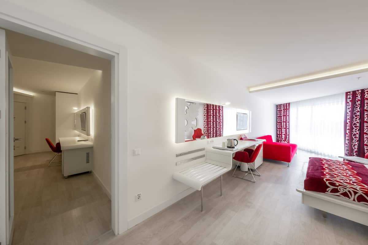 A modern hotel room with a white bed, red accent chairs, a desk with a chair, a large mirror, and a red patterned curtain.