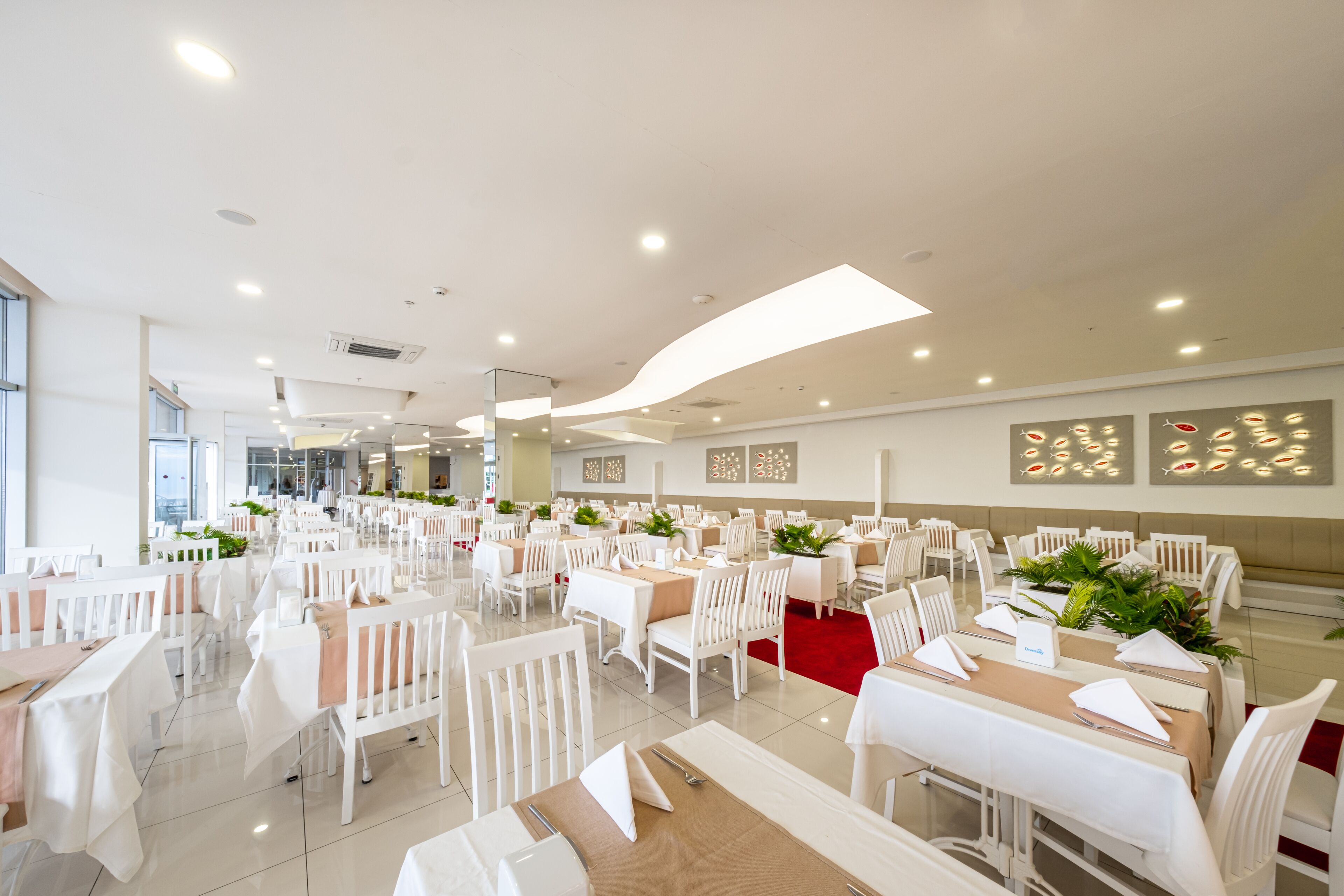 Un'ampia sala da pranzo con tavoli e sedie bianche, grandi finestre e un moderno design del soffitto.