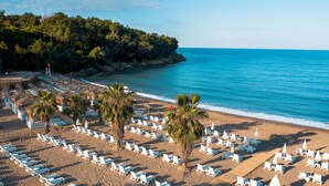 Une plage bordée de rangées de chaises longues et de parasols blancs, de palmiers et d’une côte rocheuse.