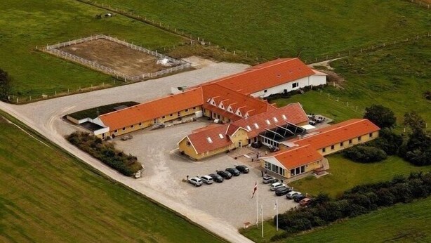 A large, rectangular building with a red roof, surrounded by green fields. It has multiple sections, including a parking area with several cars. - Hotel Højgaarden (Fjerritslev)