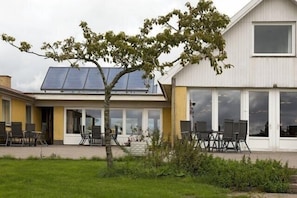 A building with a solar panel roof, a covered patio with chairs, and a tree in the foreground. - Hotel Højgaarden (Fjerritslev)