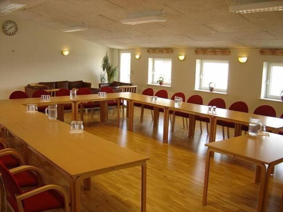 Una sala de conferencias con una mesa larga y sillas, un reloj en la pared y ventanas con plantas.