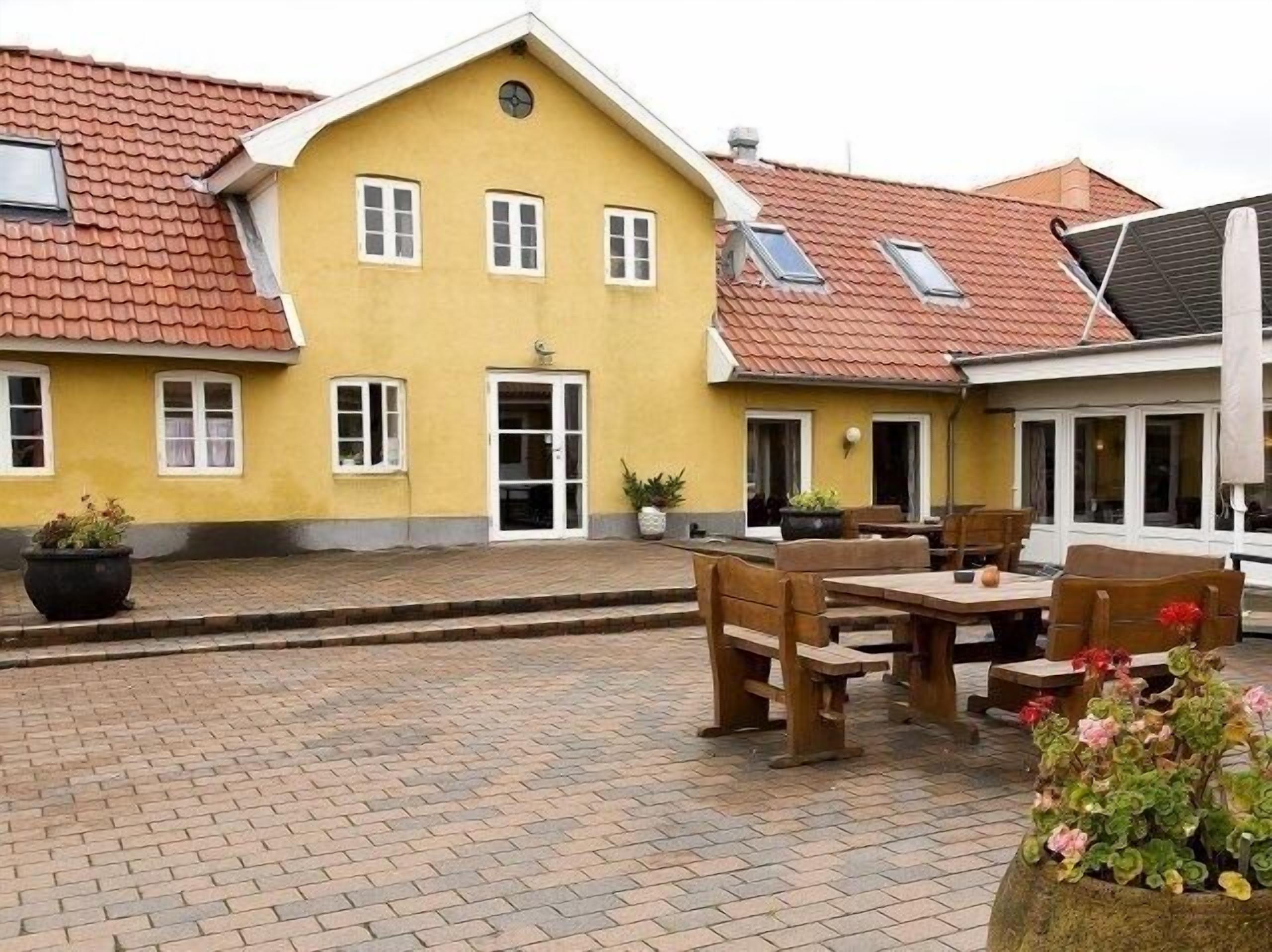Ein gelbes Gebäude mit rotem Ziegeldach, einer Haustür und einem kleinen Veranda-Bereich mit Holzmöbeln.