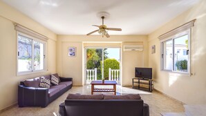 A living room with a sofa, a coffee table, a TV, and a ceiling fan. There are windows and a view of the outside. - Hadjios Valley (Mazotos)