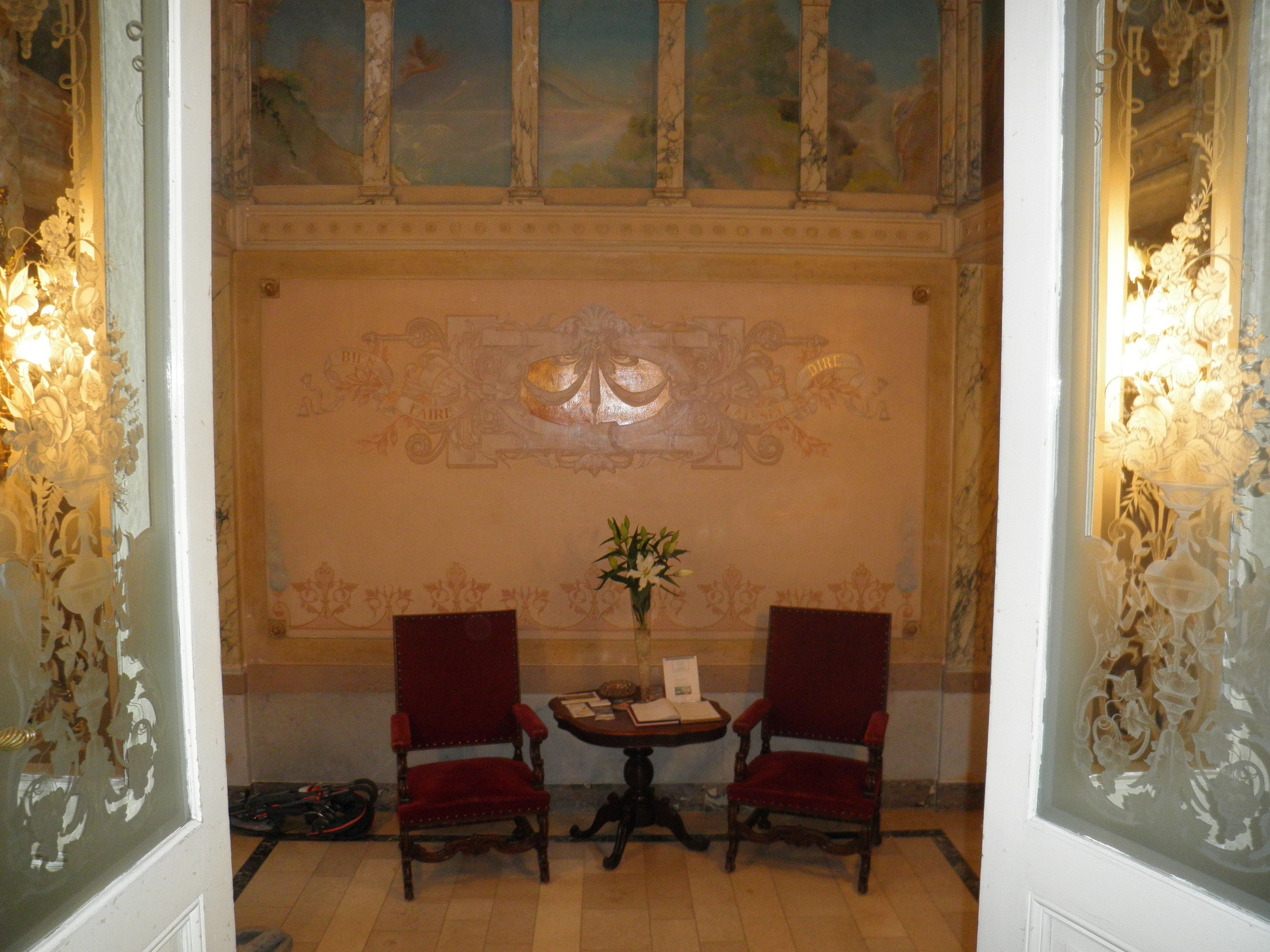 Een kamer met twee rode stoelen, een tafeltje met een bloevas en een decoratieve wand met een centraal embleem.