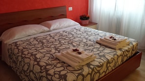 Een bed met een beddegoed met patroon, twee handdoeken met bloemen, een kleine potplant en een rode muur.