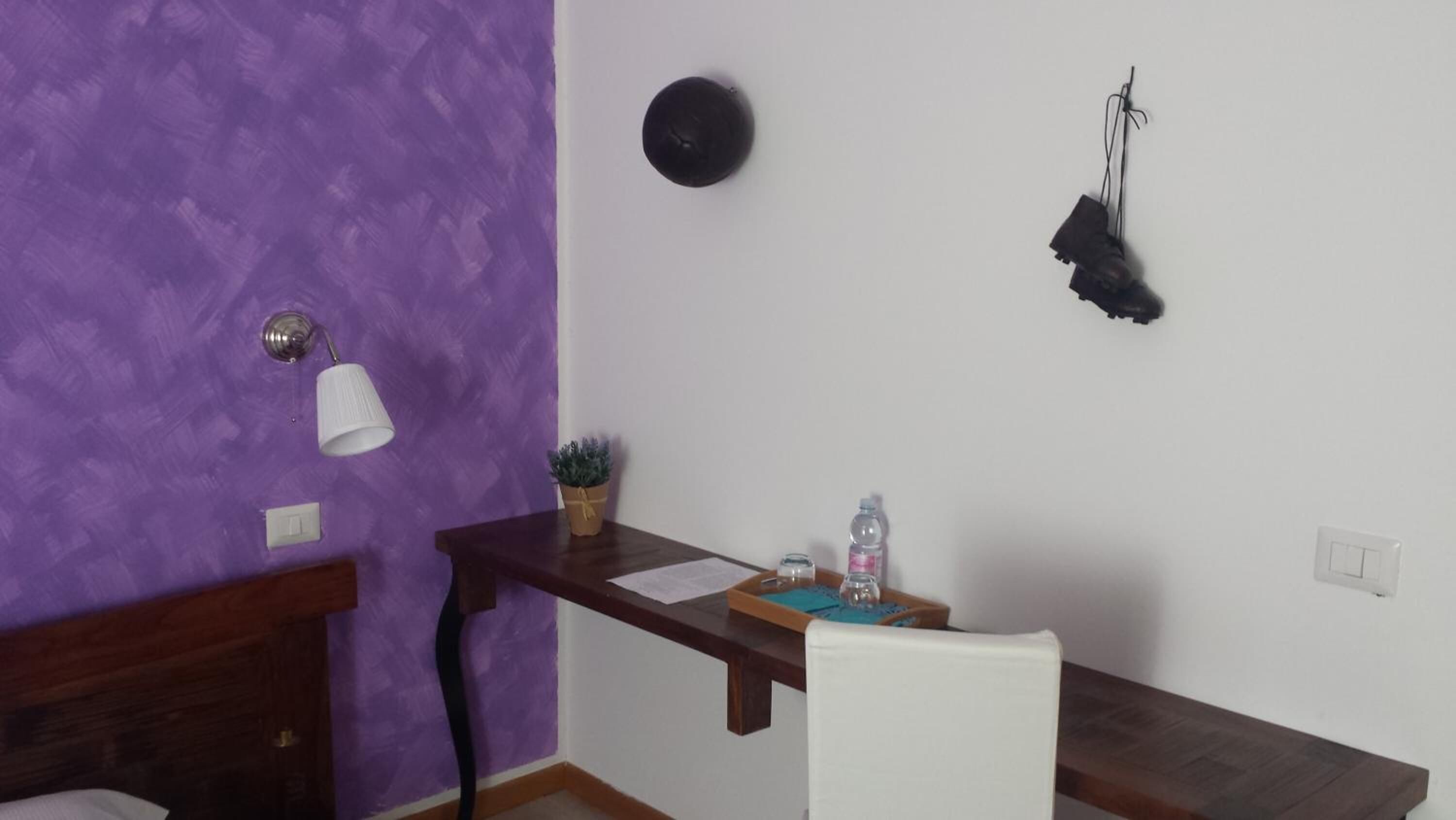 A room with a purple wall, a wooden desk, a white chair, a small potted plant, a water bottle, and a black object on the wall.