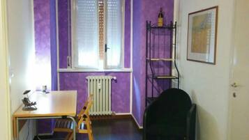 Una habitación con paredes moradas, un radiador, una ventana con persianas, un escritorio con una silla, una silla negra y un cuadro enmarcado en la pared.