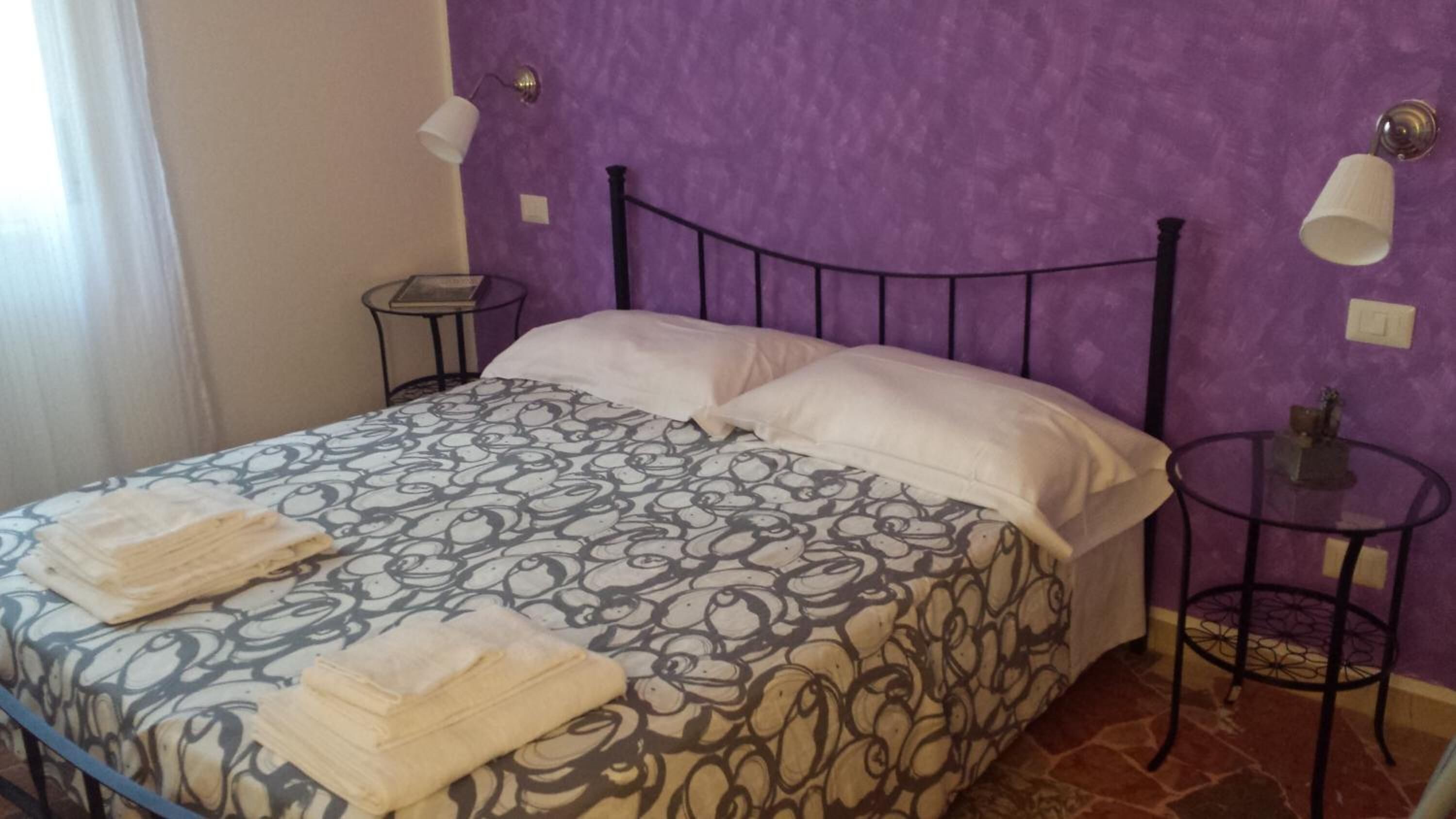 Ein Bett mit gemustertem Überwurf, zwei Kissen und gefalteten Handtüchern. Zwei Nachttische mit Lampen, eine violette Wand.