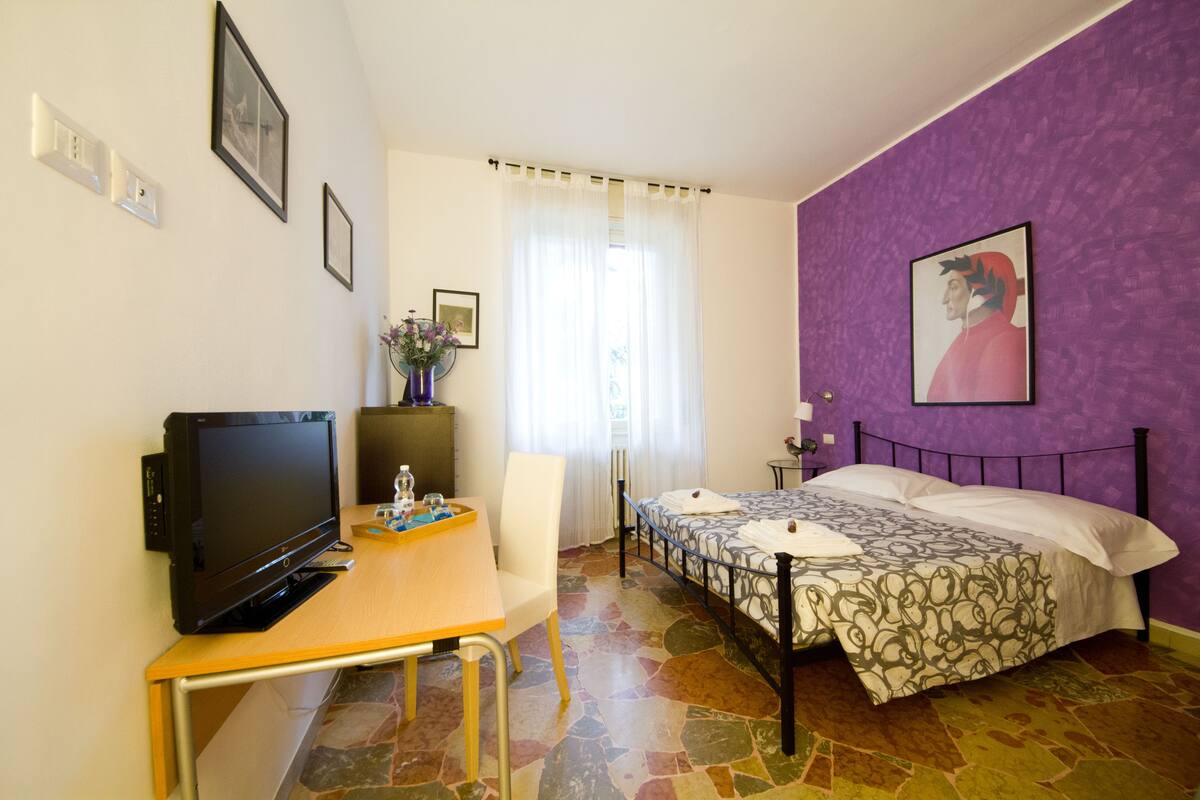 Un dormitorio con cama, escritorio con televisor, una silla y ventana con cortinas.