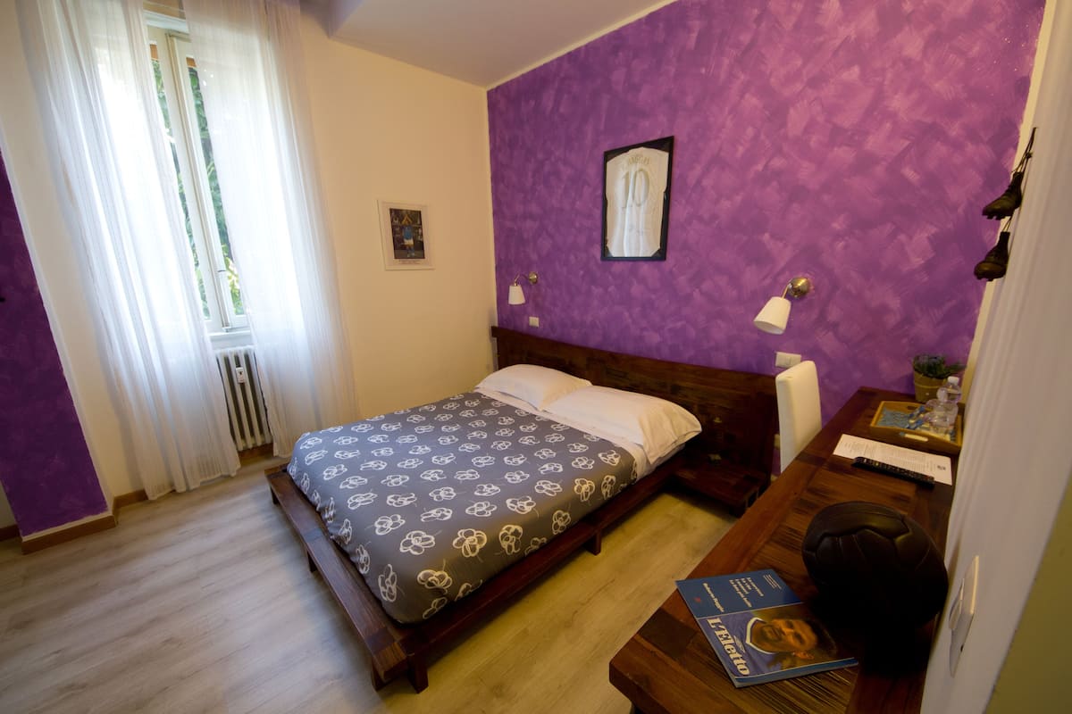 Un dormitorio con cama, escritorio y silla, ventana con cortinas y pared morada.