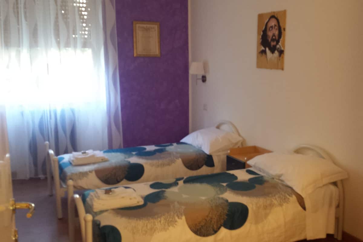 Un dormitorio con dos camas individuales, una pared morada, un cuadro enmarcado y una ventana con cortinas.