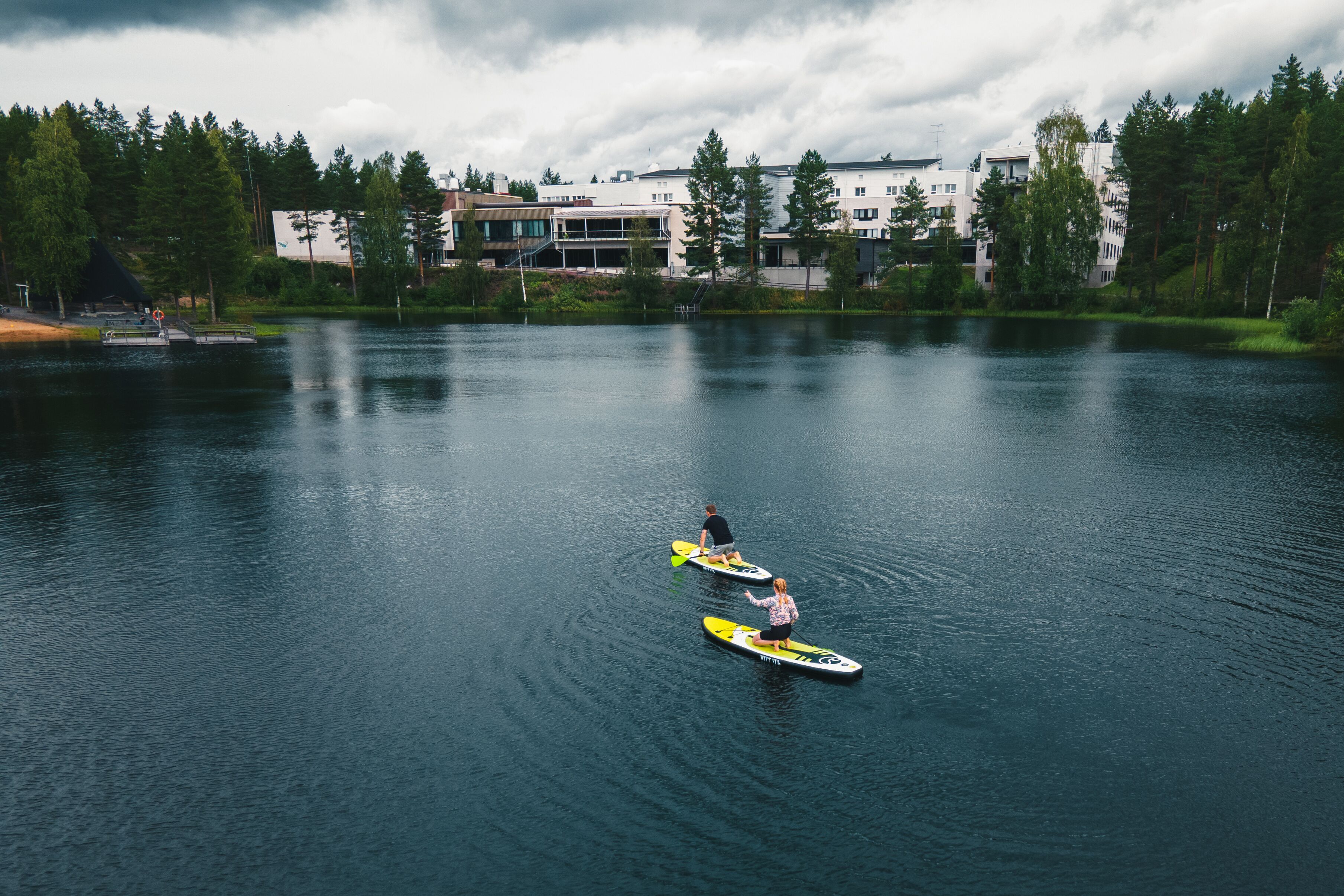 Un lago tranquilo con dos tablas de remo, un muelle y edificios al fondo.
