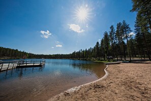 A serene lakeside scene with a wooden dock extending into the water, surrounded by dense forest and clear skies.