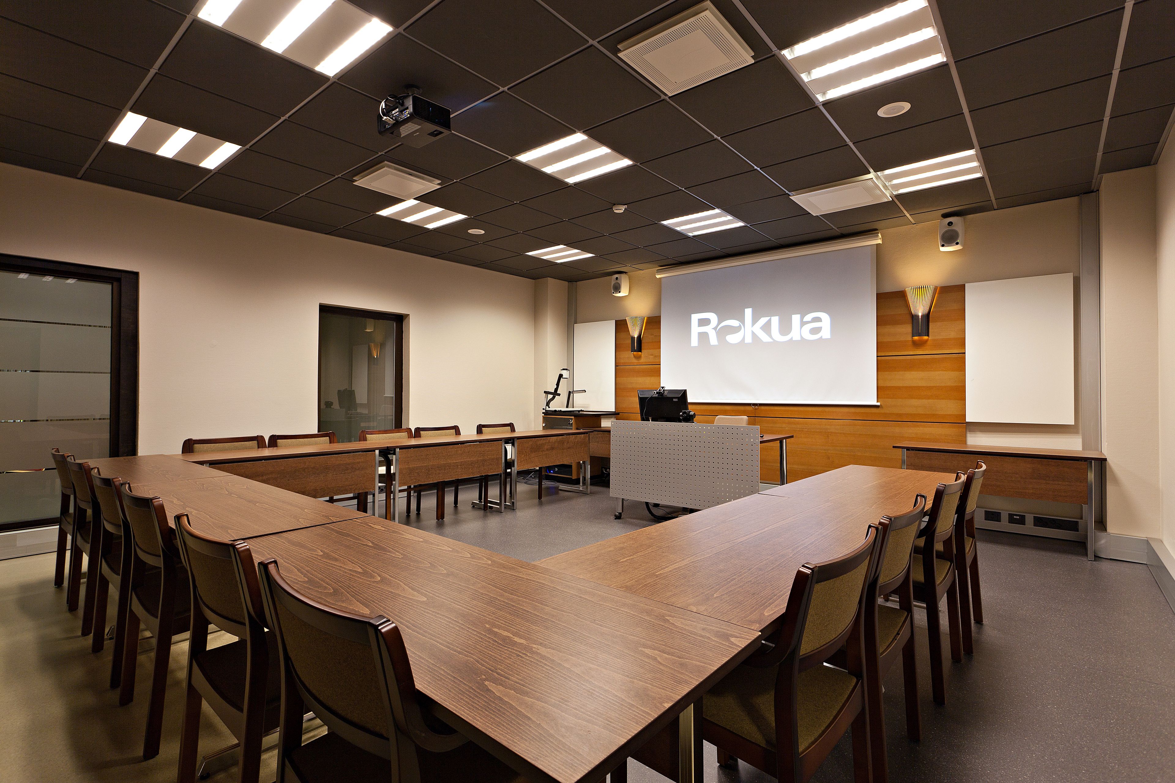 Un salón de conferencias con una gran mesa de madera, sillas, una pantalla de proyector y una pantalla de presentación que muestra la palabra "Rokuia".