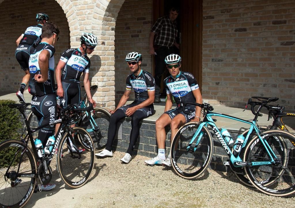 A group of cyclists in cycling attire seated and standing near bicycles, with a brick building in the background.