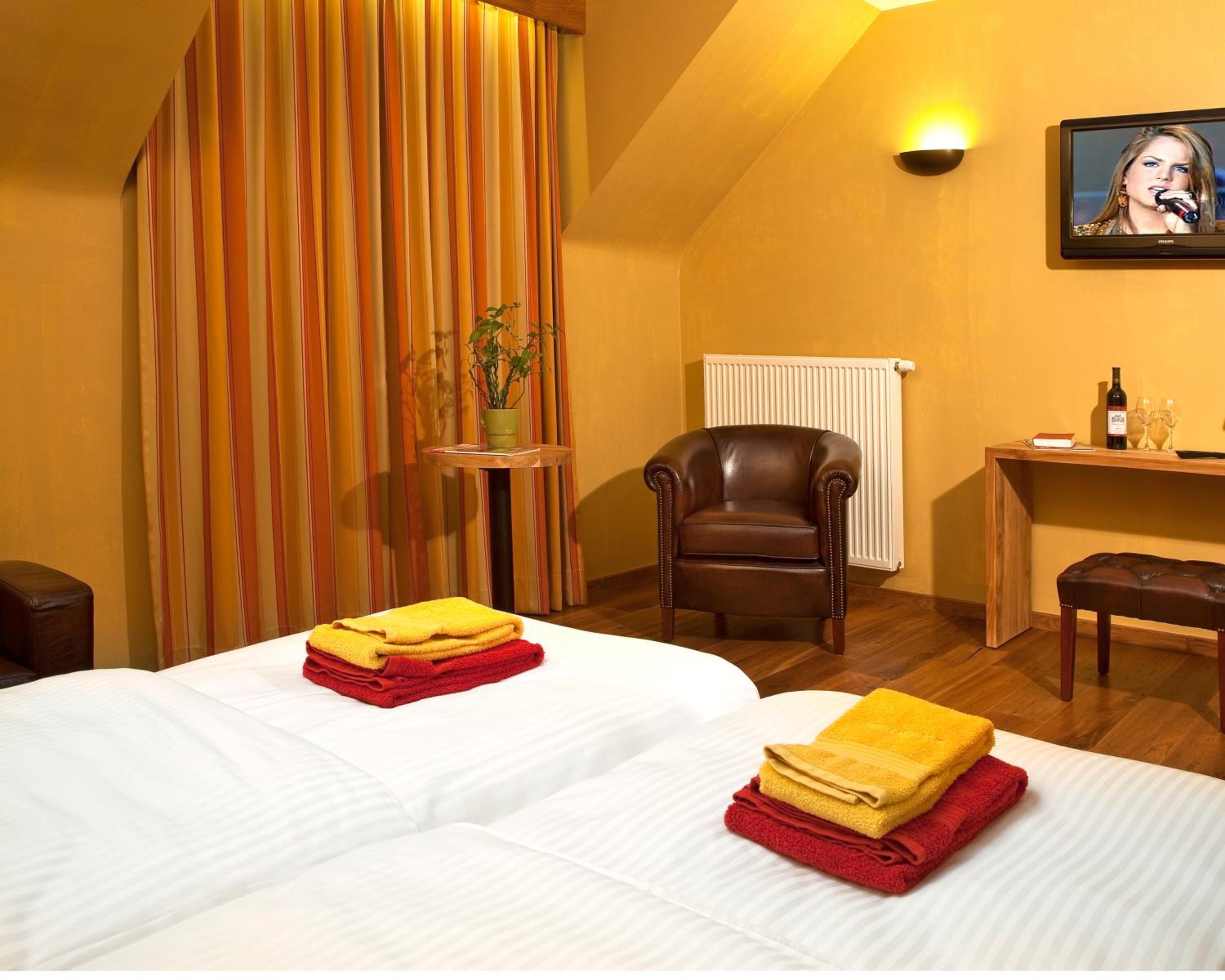 Habitación de hotel con dos camas, un sillón de cuero marrón, una mesita con una planta, un radiador y un escritorio con una botella y vasos.