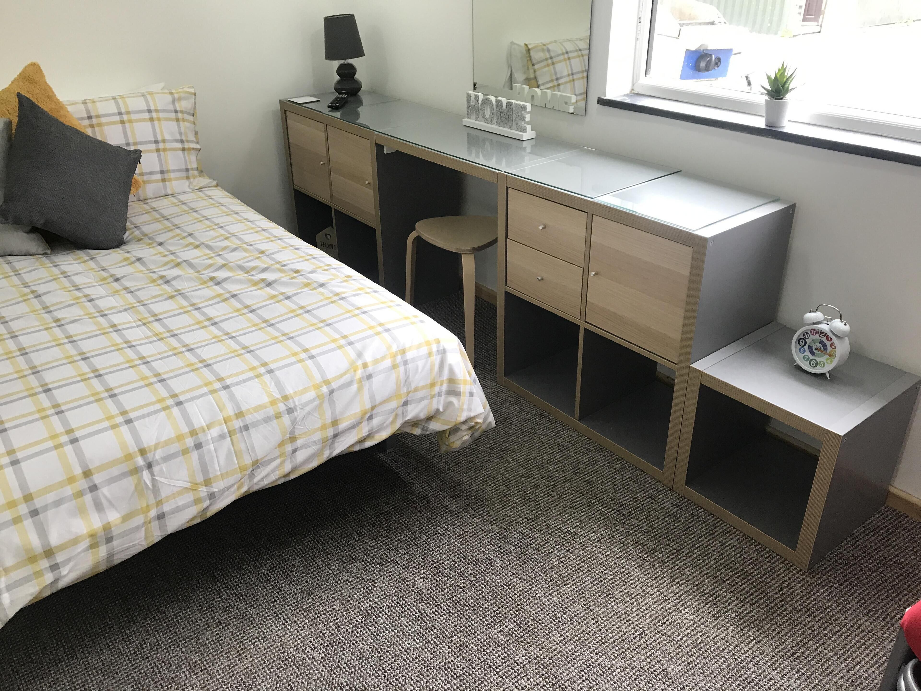 A bedroom with a bed, a desk, a chair, a small table, and a nightstand.