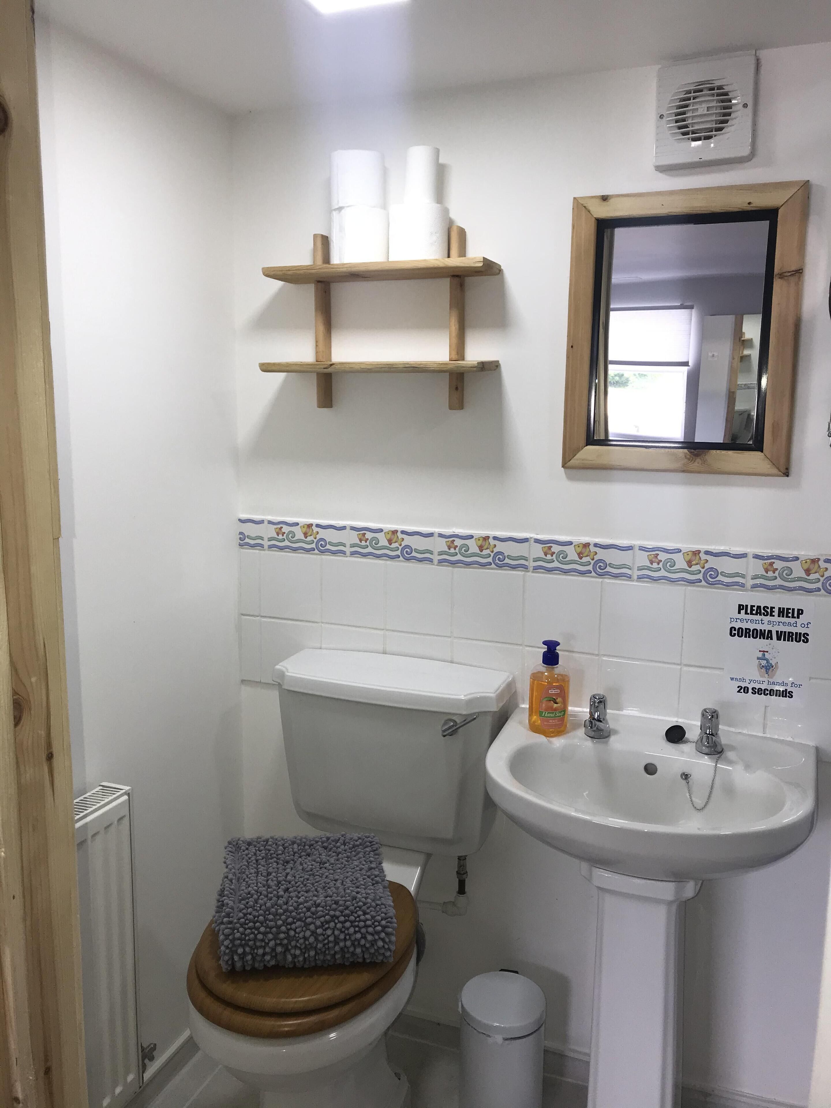 Un baño pequeño con inodoro, lavamanos y espejo. Hay rollos de papel higiénico en un estante sobre el inodoro.